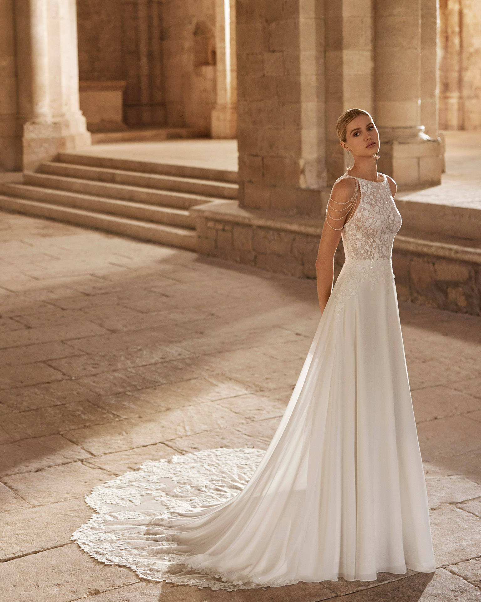 Νυφικό φόρεμα από πολύ λεπτή ανάλαφρη georgette, σε Α-line γραμμή από την νυφική συλλογή Rosa Clara. Δαντελένιο μπούστο, κεντημένο με χαντράκι με λάμψη, κλειστό μπροστά στο μπούστο λαιμόκοψη bateau και τιράντες φτιαγμένες από χάντρες σε όλη την ανοιχτή πλάτη. Ένα μοναδικό νυφικό φόρεμα από την Rosa Clará για νύφες ξεχωριστές.